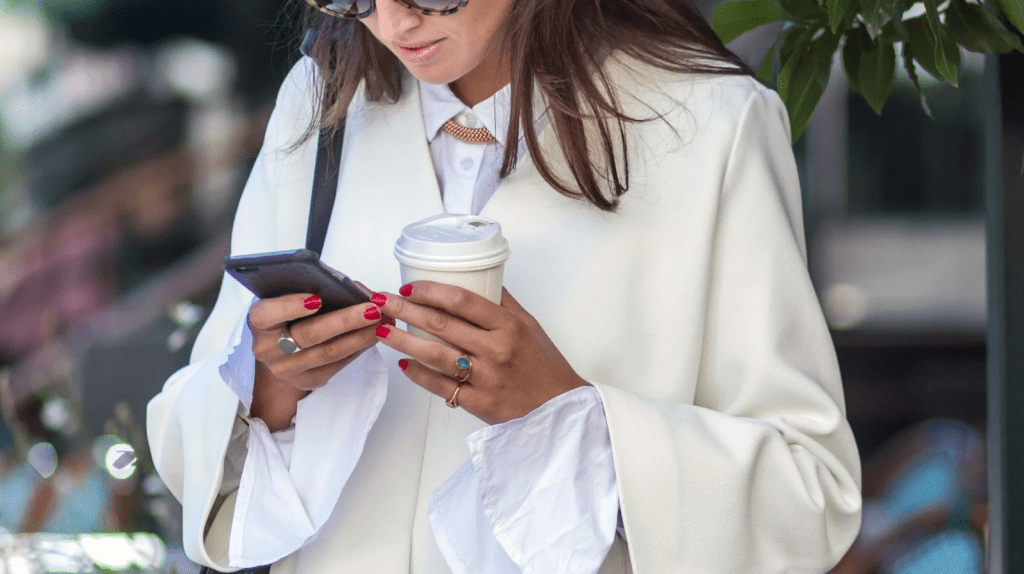 A professionally dressed woman holds a coffee in one hand and her phone in the other. She looks down at her phone.