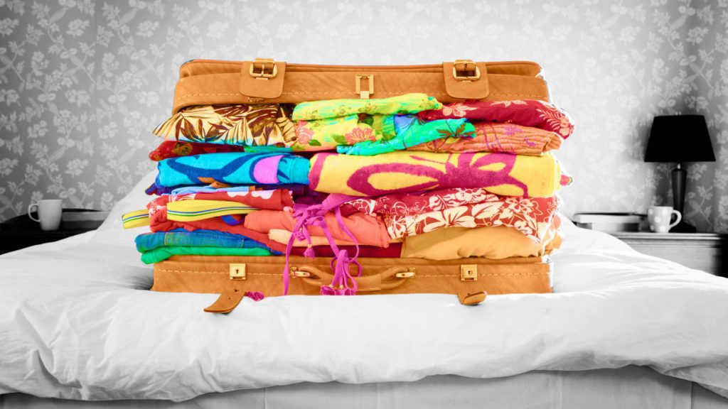 A suitcase sits on a bed in a black a white room. The suitcase has piles of colorful vacation clothes, which will likely prevent it from closing.