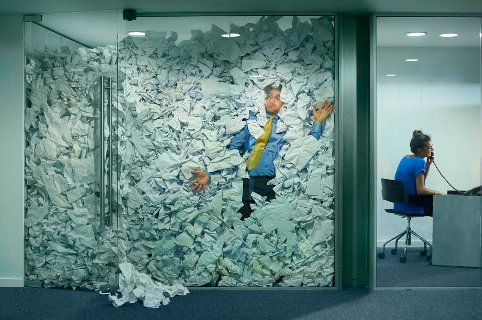 A man is pressed against the glass wall of his cubicle, which is entirely filled by crumbled paper. He looks stressed, while the woman in the cubicle next to him smiles during a phone call.
