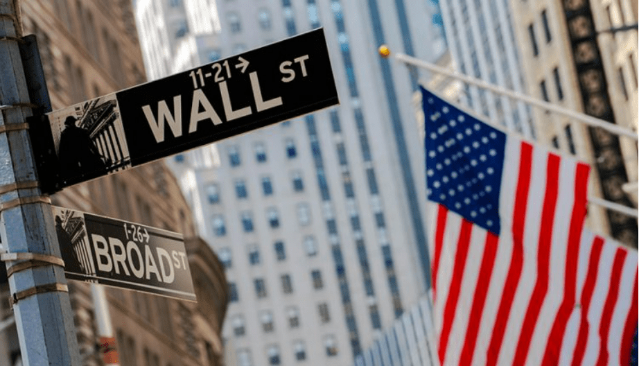A photo of a Wall Street sign in New York city on the left side of the street. The American Flag hangs on the right hand side also in focus. There is a tall office building in the background.