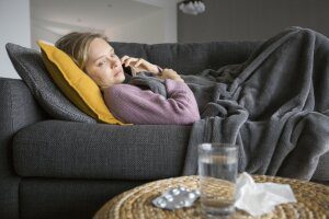 A woman lays on her couch while on the phone. She is covered with a blanket and look toward her table which has water and a crumbled tissue.